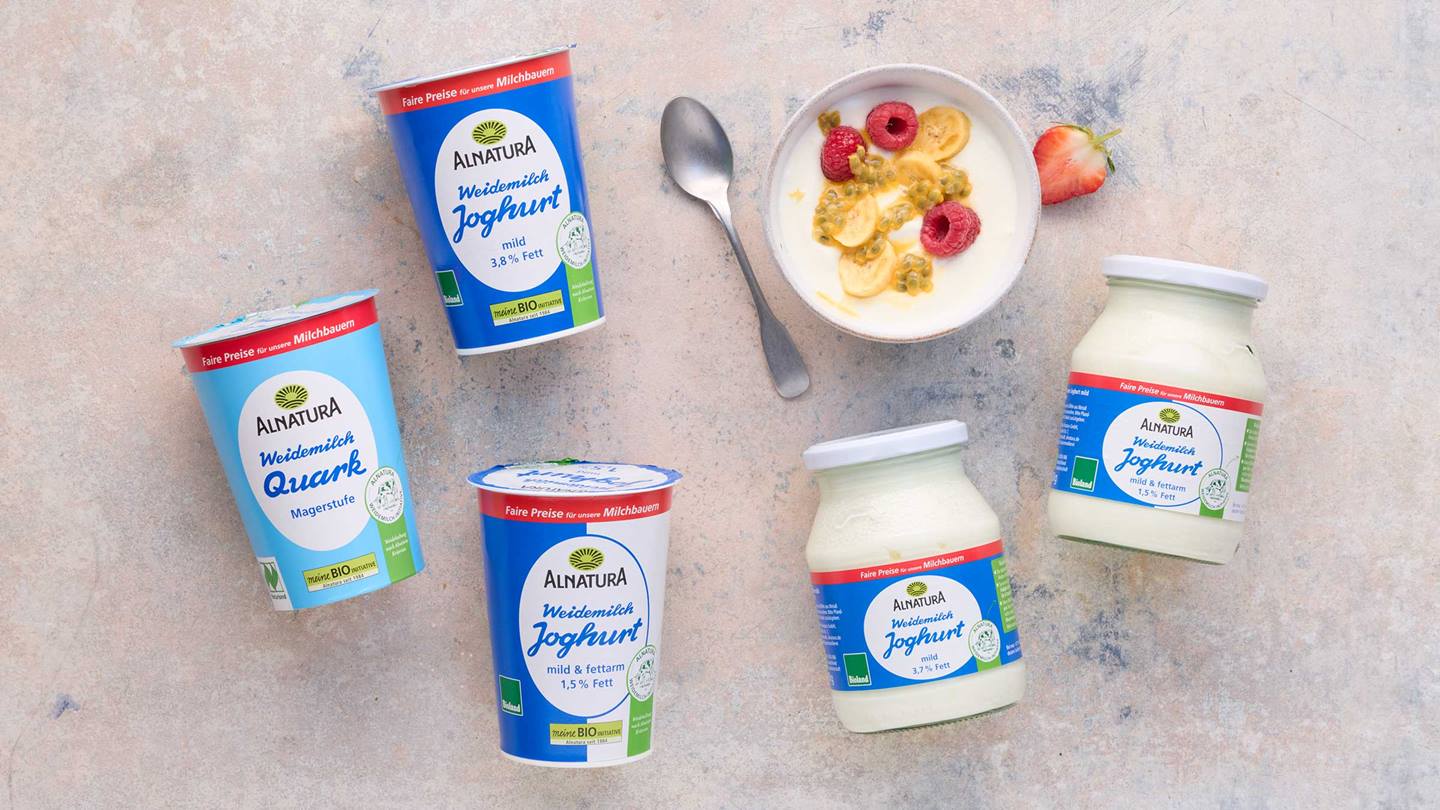 Das Bild zeigt verschiedene Produkte der Marke Alnatura: Joghurt (mild, 3,8% Fett; mild & fettarm, 1,5% Fett) und Magerquark. Die Produkte sind auf einem hellen Untergrund arrangiert, neben einer Schale Joghurt mit Früchten (Himbeeren, Banane, Passionsfrucht) und einem Löffel. Das Bild soll die Produkte präsentieren.