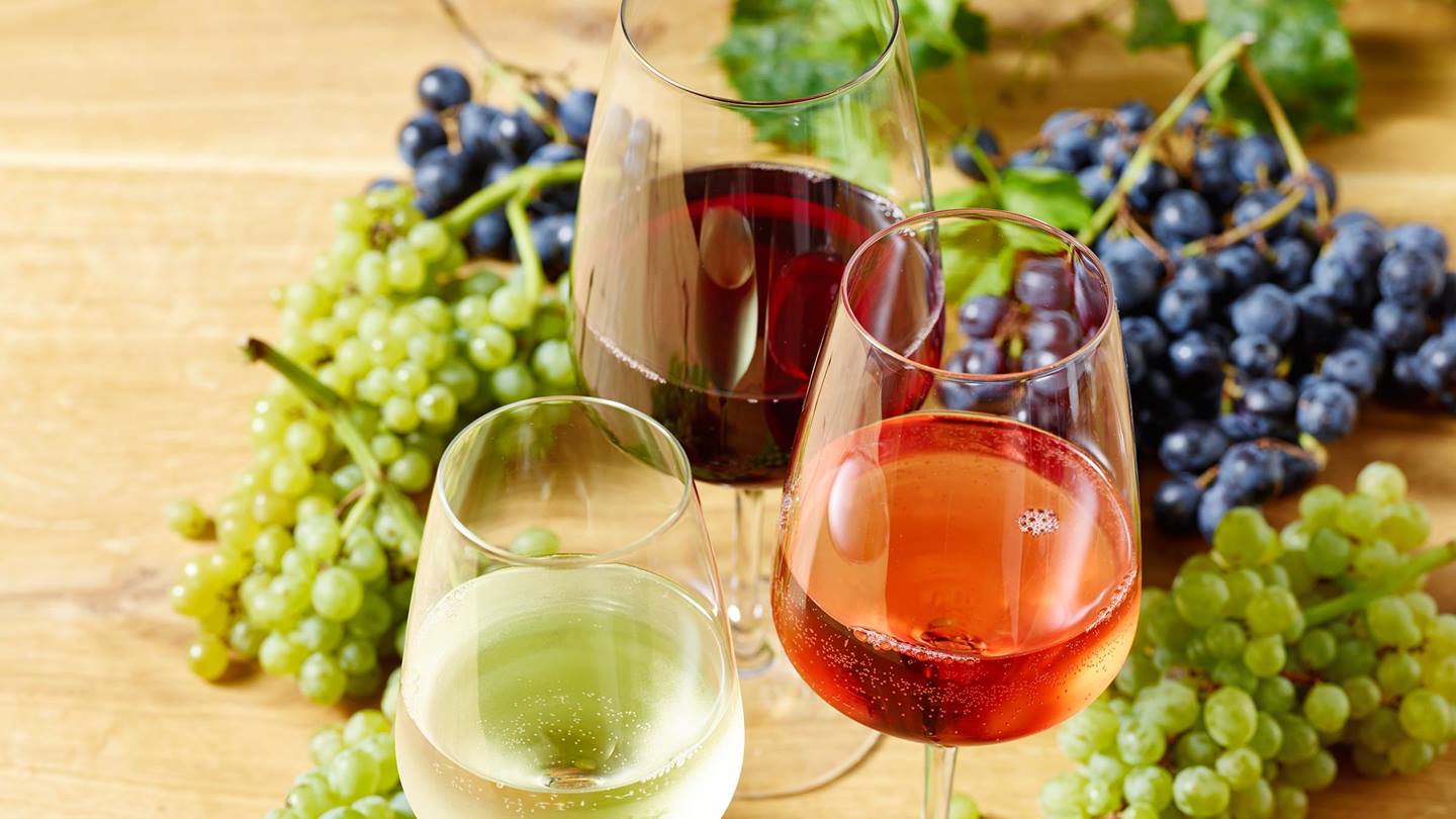 Drei Weingläser, befüllt mit unterschiedlichen Weinsorten. Ein Rotwein, ein Weißwein und ei Rosé. Darum sind blaue und helle Weintrauben dekoriert. Alles befindet sich auf einem Holzuntergrund. Im Hintergrund sind noch Weinblätter zu sehen.